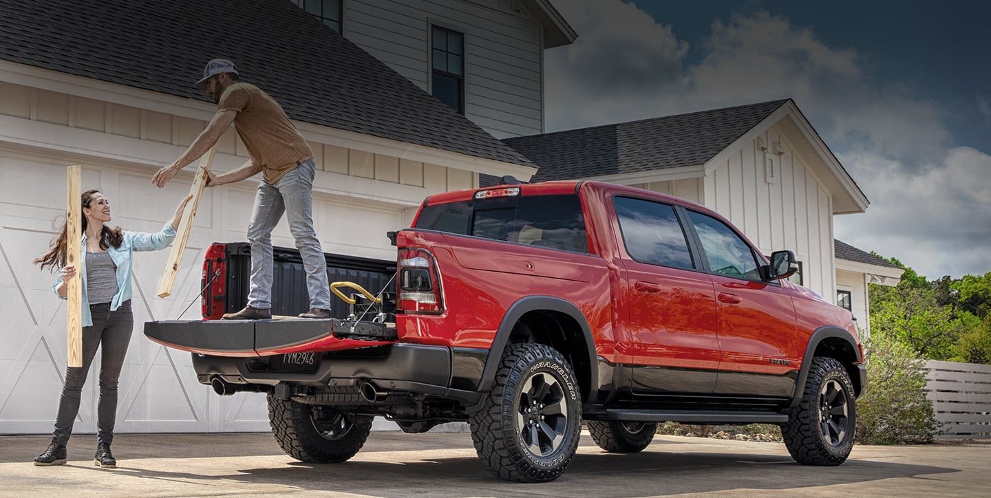 A man stands on the lowered tailgate of a 2020 Ram 1500 as a woman hands him lumber to be loaded into the pickup bed.
