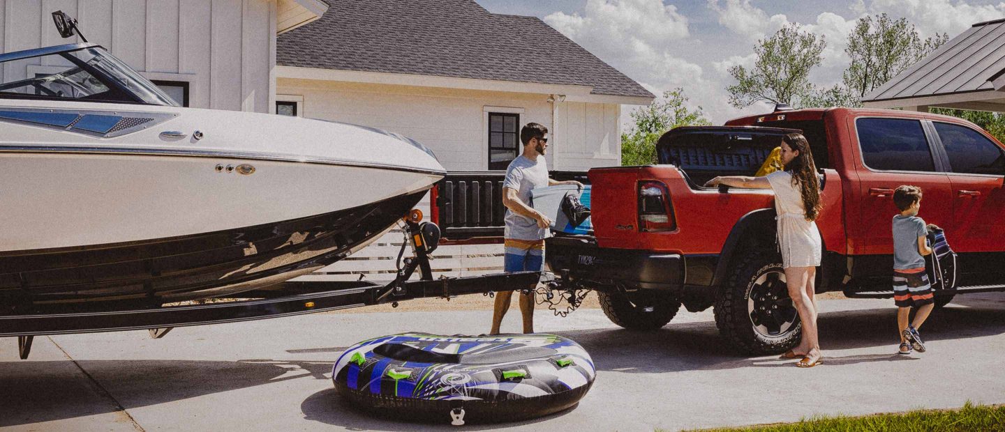 A man loading a wakeboard through the open tailgate door of a 2022 Ram 1500 with an attached boat trailer.
